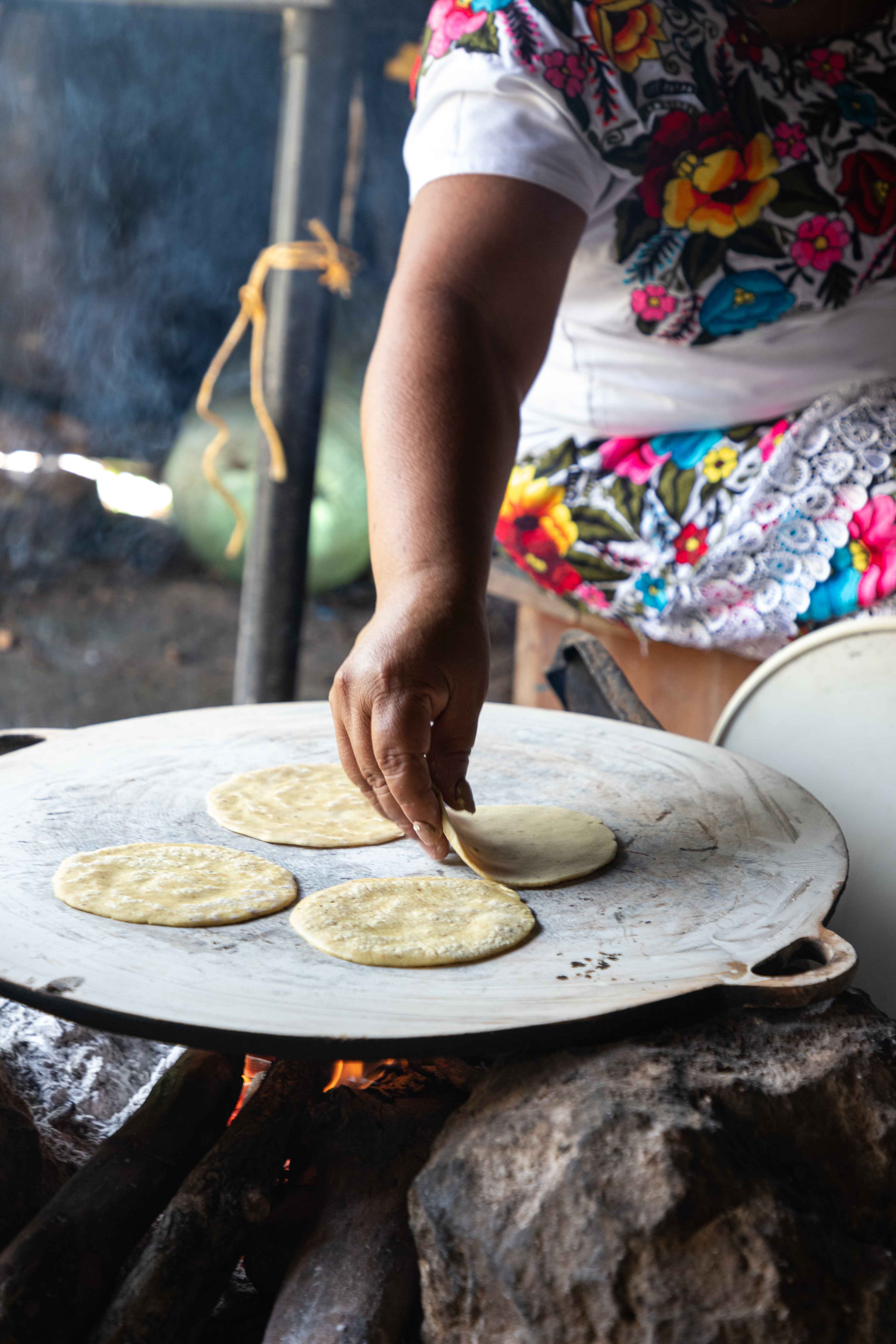 A cook in an embroidered huipil turning corn tortillas on a comal over a wood fire
